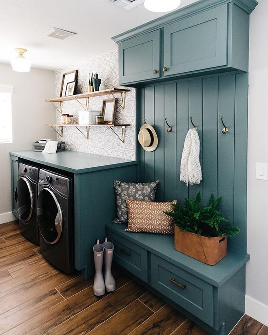 A Boho laundry room with teal cabinetry, a washer and dryer, wood shelves, patterned wallpaper, hooks with accessories, a bench with pillows, a plant, and wood-look flooring adds flair to your boho laundry decor.