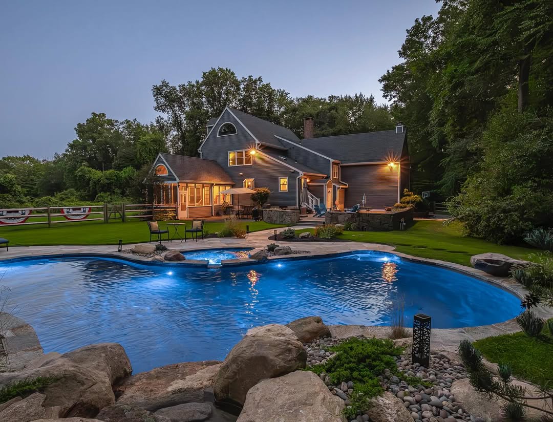 A suburban house with outdoor lights is seen at dusk, featuring a landscaped backyard with a curved swimming pool—perfect for those seeking cool pool ideas—with patio seating and surrounding trees.