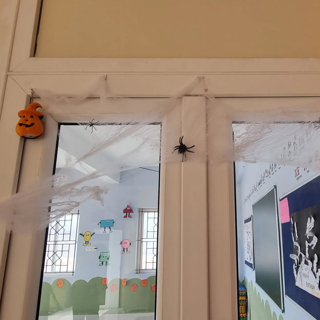 A classroom door adorned with fake spider webs, plastic spiders, and a pumpkin face ornament offers fun Halloween window decoration ideas; festive decor and student drawings are visible inside.