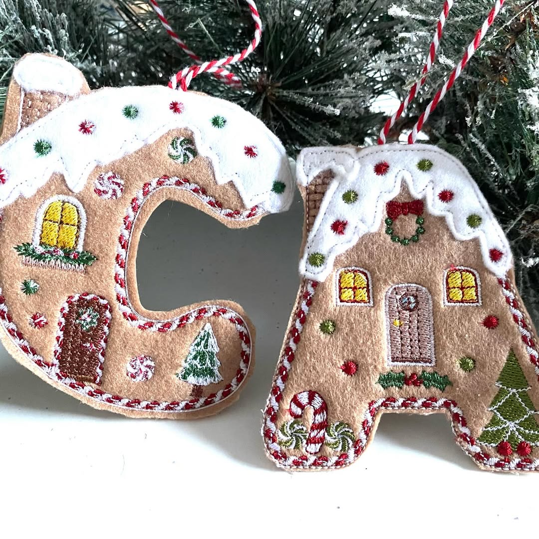 Two felt Christmas ornaments shaped like the letters C and A, decorated with embroidered gingerbread house designs, add a charming touch of Gingerbread Christmas decor as they hang in front of snowy pine branches.