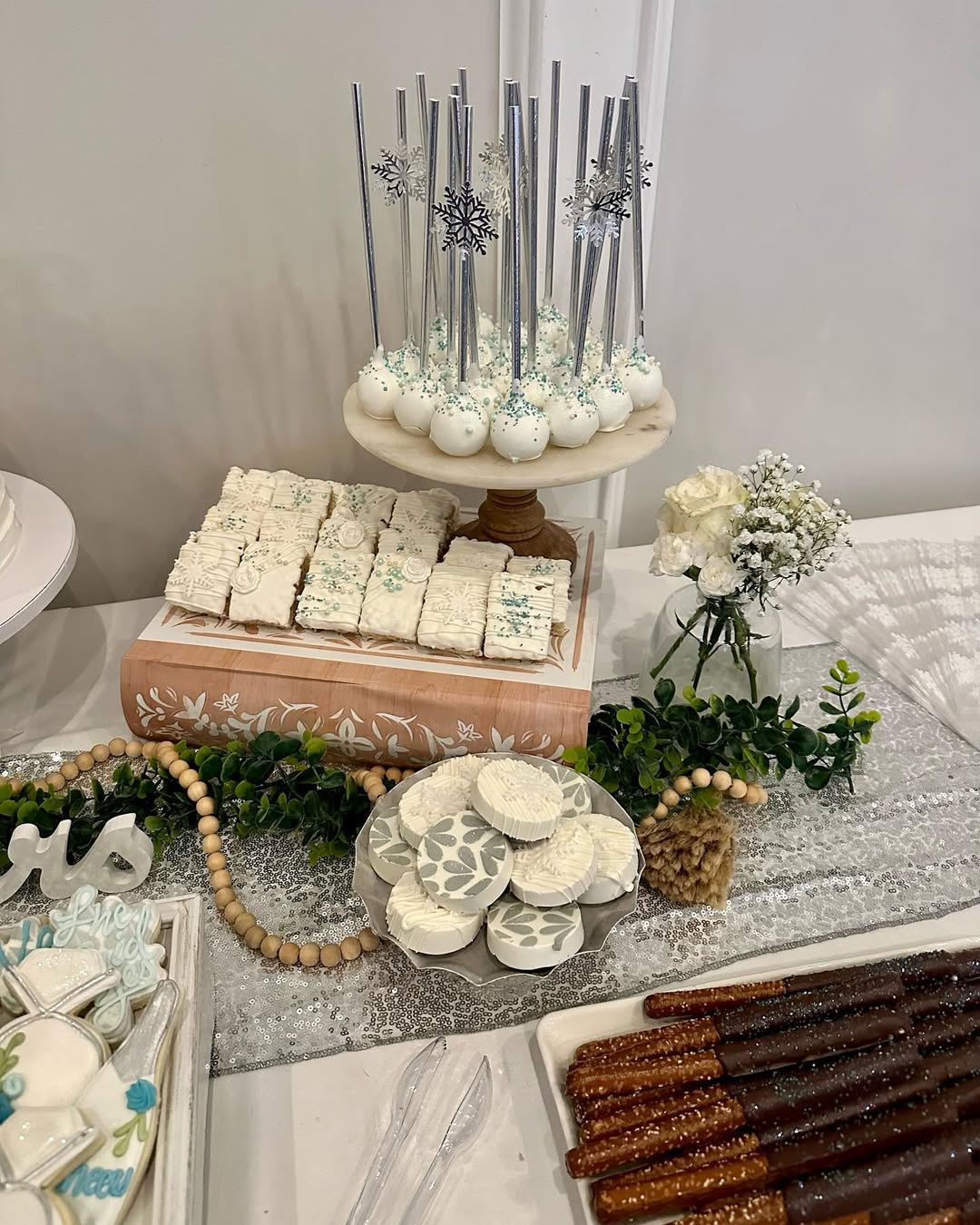 A dessert table for any occasion features white treats like cake pops with silver sticks, frosted squares, round cookies, and chocolate-covered pretzel rods, all beautifully decorated with greenery and white flowers—perfect for dessert table ideas.