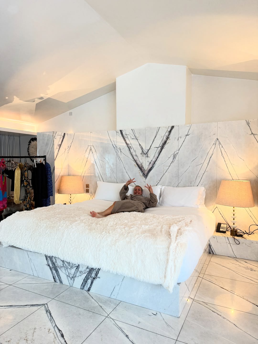 A person in a robe lies on a large marble bed in a spacious, modern women bedroom with marble floors, two lamps, and clothing racks in the background—perfect for those seeking elegant women bedroom ideas.
