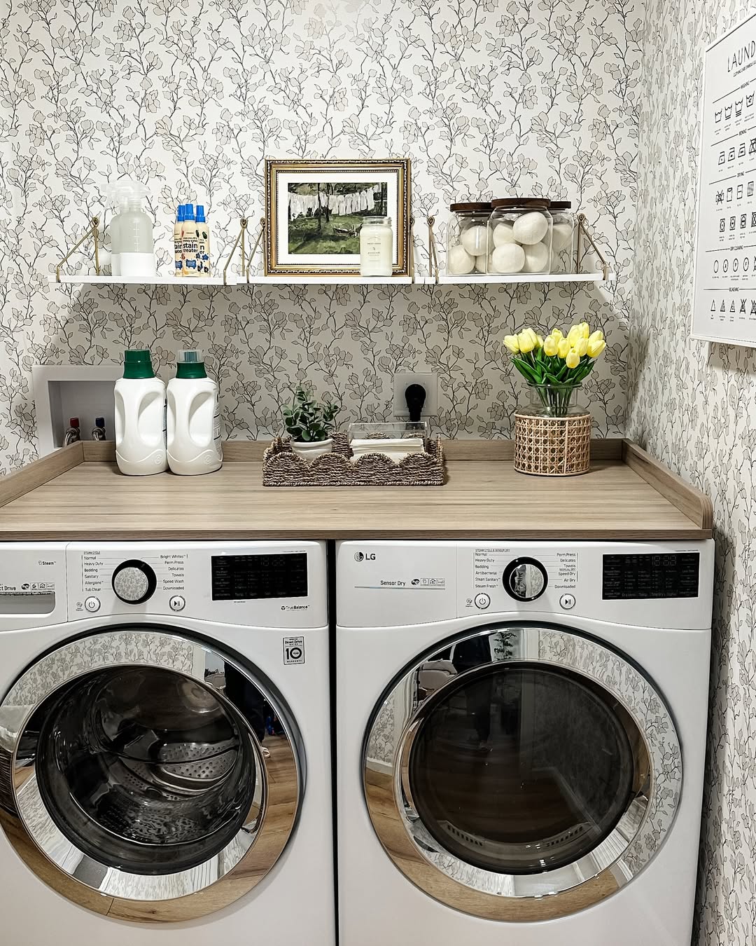 A boho laundry room featuring a washer and dryer, wooden countertop, detergent bottles, a tray with decor, a vase of yellow tulips, and shelves holding jars, framed art, and cleaning supplies offers fresh laundry room ideas.