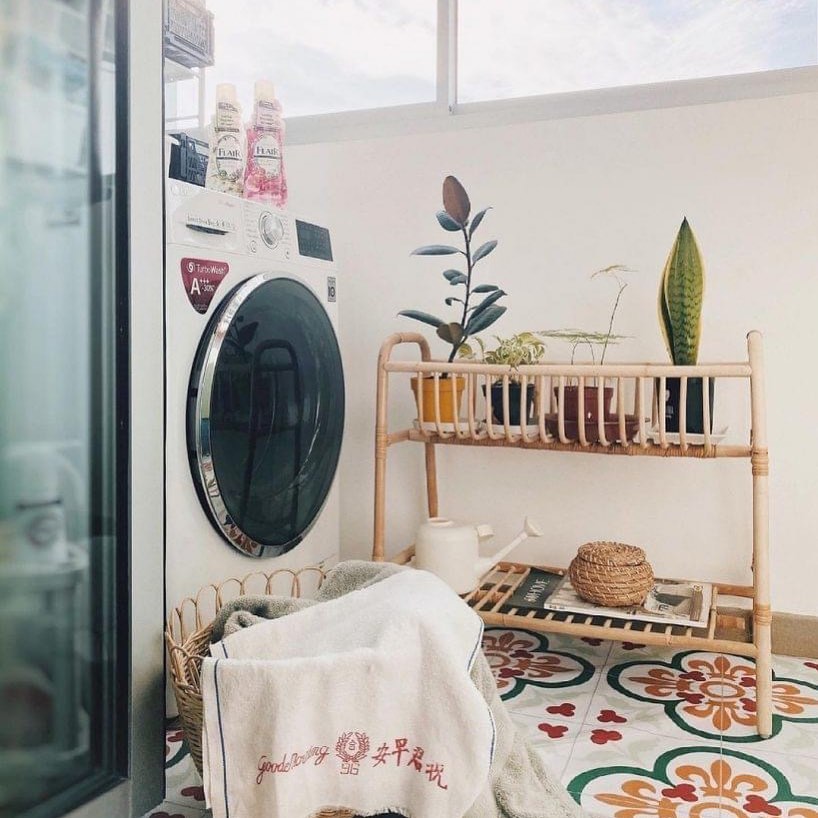 A Boho laundry room featuring a front-loading washing machine, potted plants on a bamboo shelf, a watering can, and a folded towel resting on colorful patterned floor tiles—perfect inspiration for boho decor and laundry room ideas.