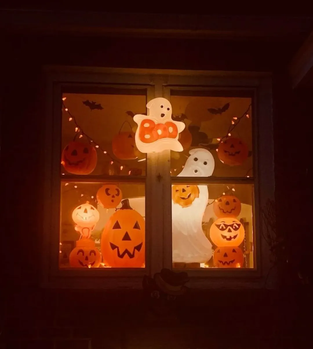A window decorated with glowing jack-o’-lanterns, ghost figures, and orange string lights offers inspiring Halloween window decoration ideas for a festive display at night.