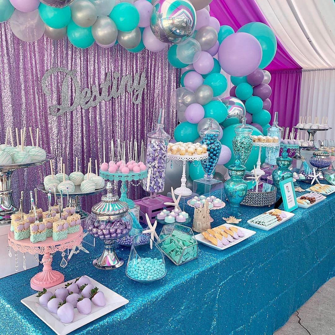 A birthday dessert table decorated with teal, purple, and silver balloons, a glittery backdrop, assorted party desserts, and a sign reading