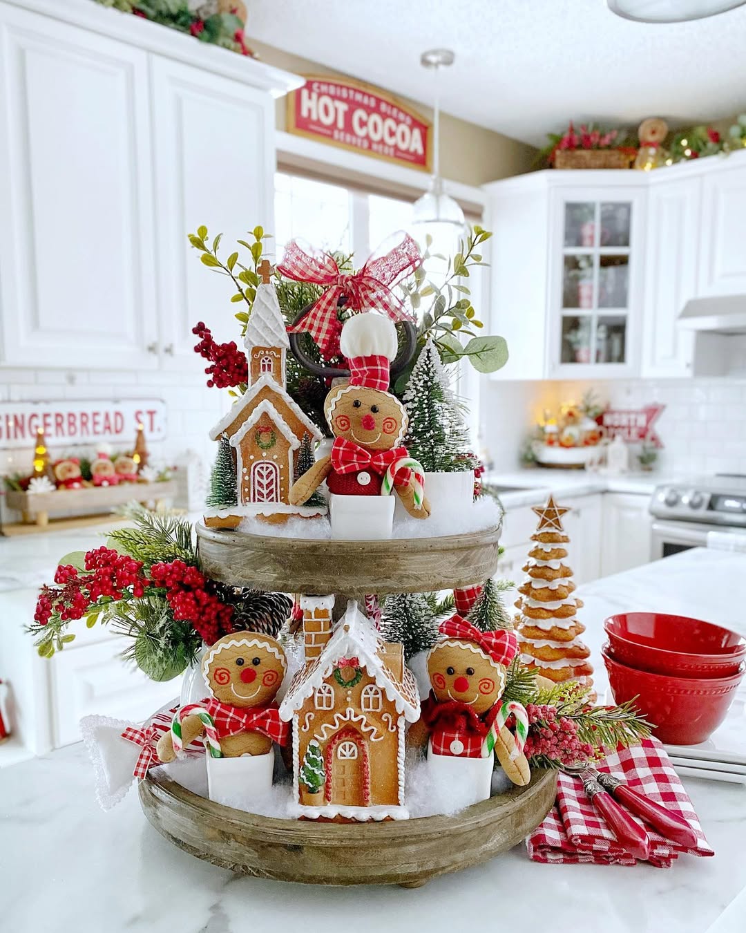 A tiered wooden tray displays gingerbread houses, gingerbread figures, festive greenery, candy canes, and Gingerbread Christmas decor on a white kitchen counter.