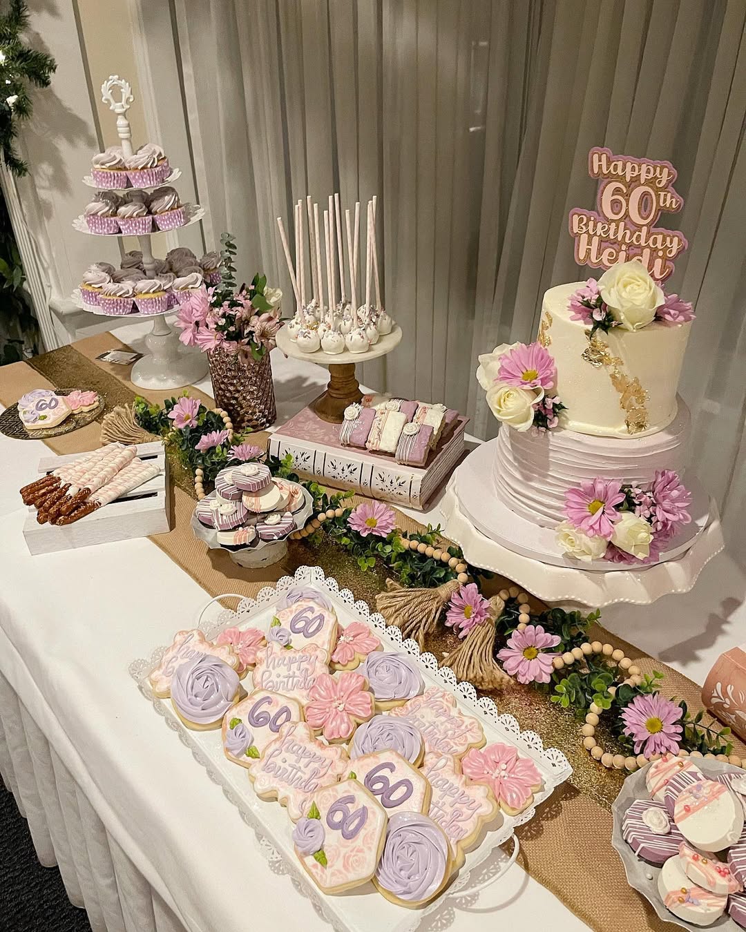 A decorated dessert table for a 60th birthday features pretty desserts like cake, cookies, cupcakes, and cake pops, with floral arrangements in pink, purple, and white tones—perfect for party desserts inspiration.