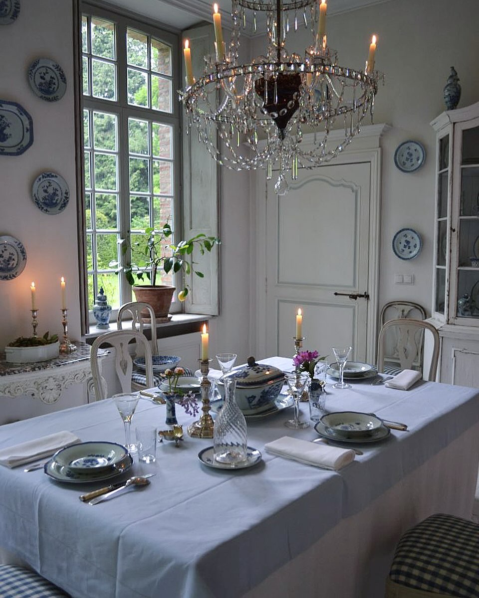 A dining table set for four with white linens, blue and white dishes, glassware, candles, and a crystal chandelier offers timeless table decor in a light-filled, elegant room with vintage charm—perfect for dining table inspiration.