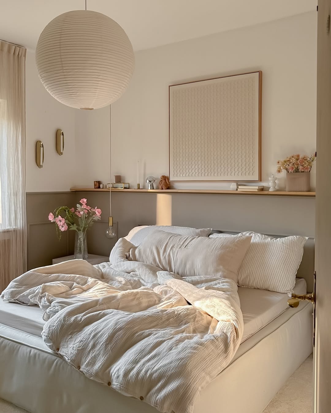 A neatly made bed with beige bedding is in a softly lit small bedroom, featuring a round paper lamp, flowers in a vase, and minimalist decor on a shelf above the headboard—perfect inspiration for small bedroom layout ideas.