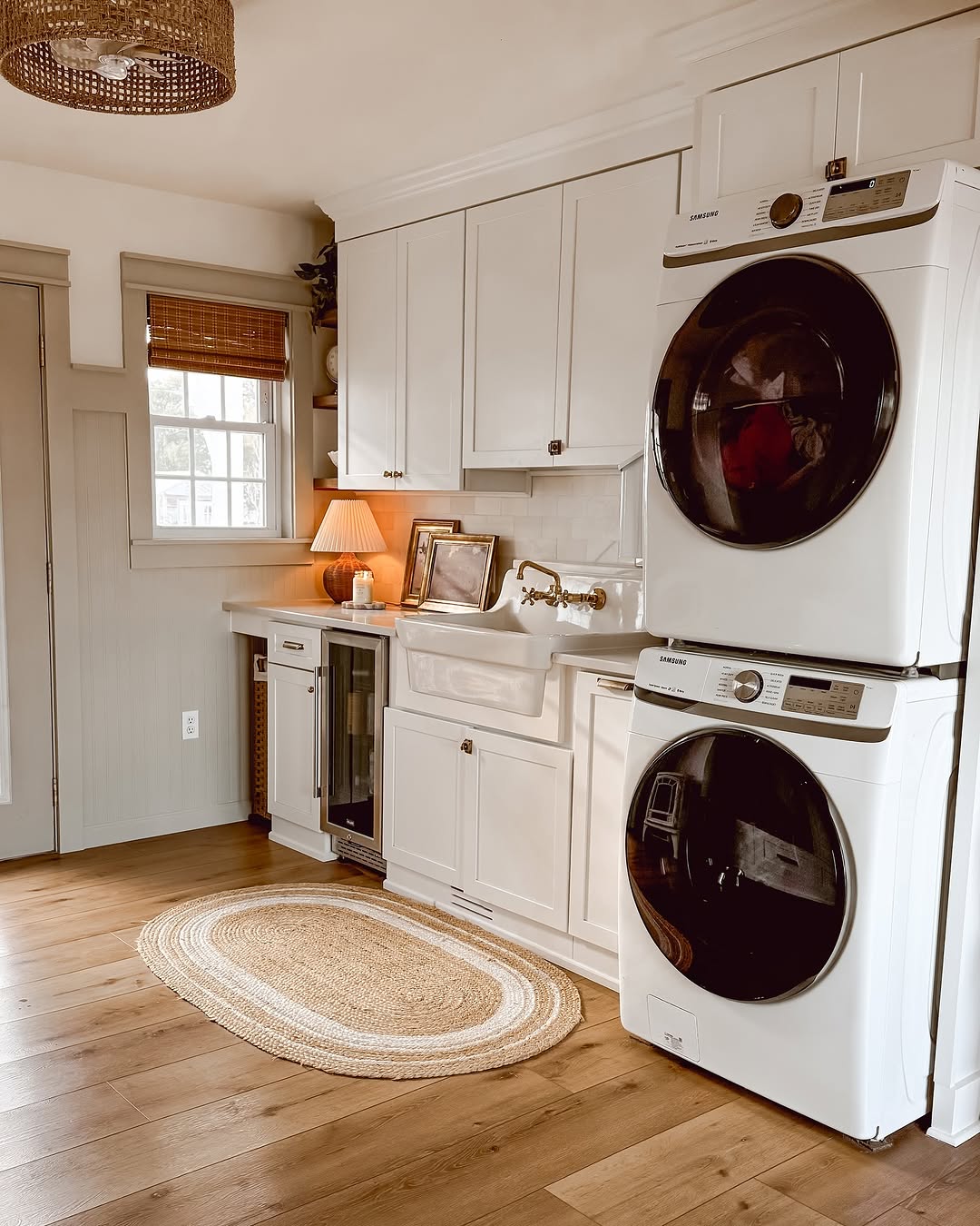 Cozy small laundry room with stacked washer and dryer, white cabinets, wooden floor, and a circular rug. A sink, framed pictures, lamp, and window with blinds complete the charming space.
