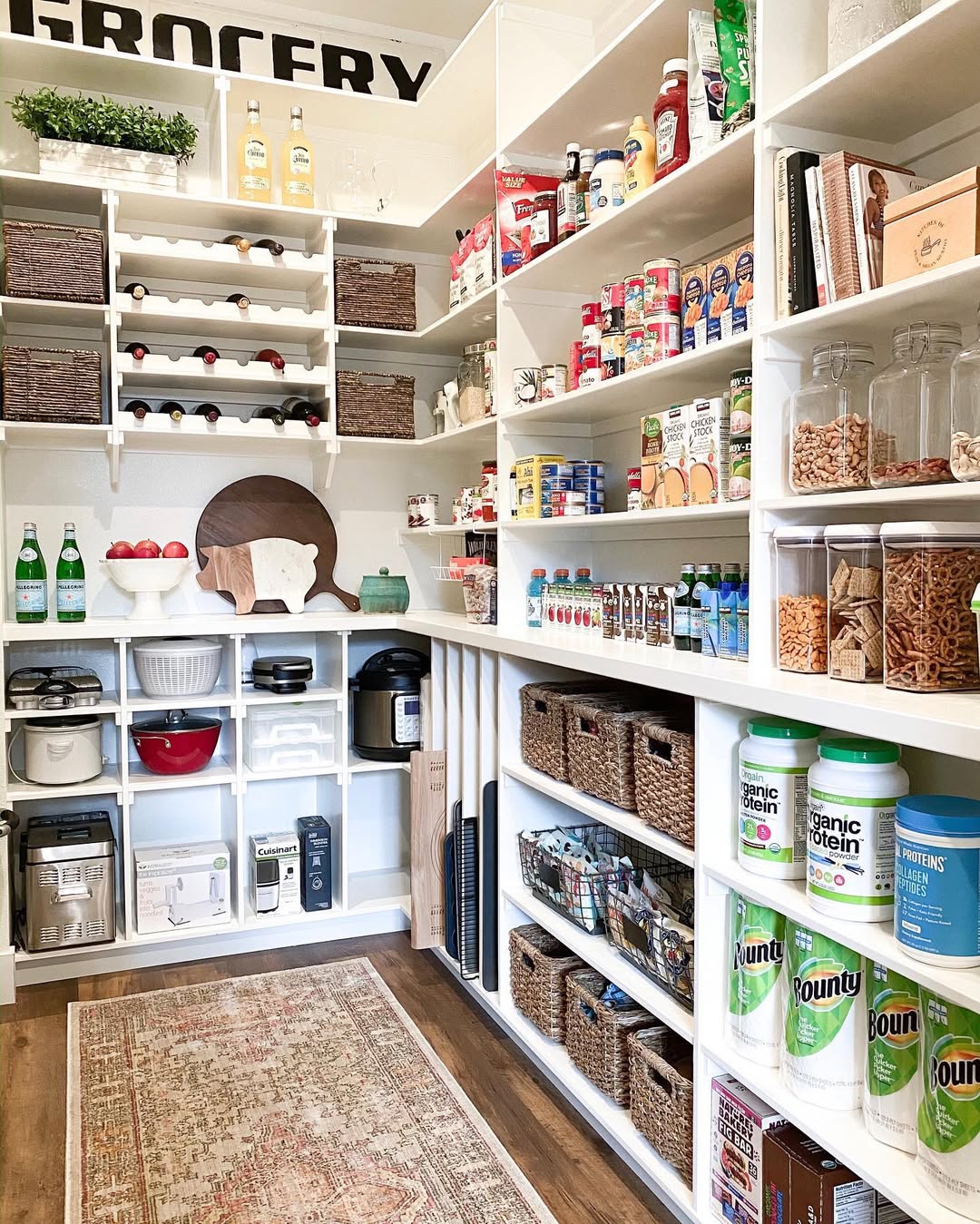 A neatly organized pantry showcases excellent pantry organization, with shelves holding baskets, jars of dry goods, canned foods, kitchen appliances, and large containers of protein powder and paper towels.