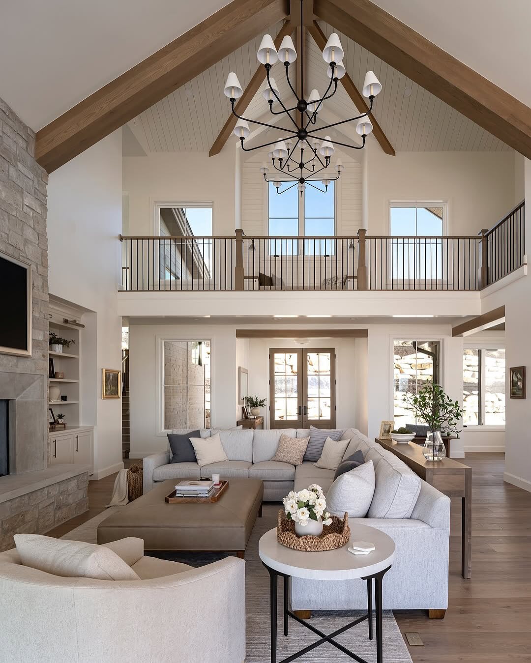 Spacious organic modern living room with vaulted ceiling, exposed wooden beams, sectional sofa, stone fireplace, chandelier, and an upstairs balcony overlooking large windows—perfect for inspiring new living room ideas.