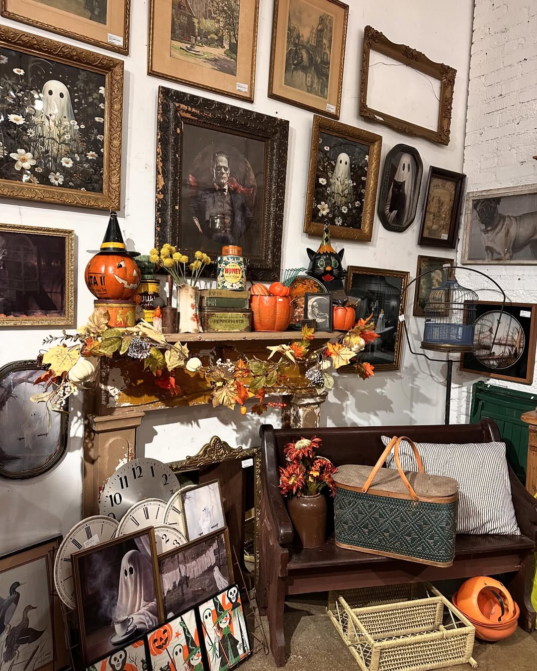 A wall displays framed vintage Halloween art above a shelf with pumpkins, fall decor, and a bench holding a picnic basket; vintage clocks and ghost art add to the nostalgic Halloween decor on the floor.