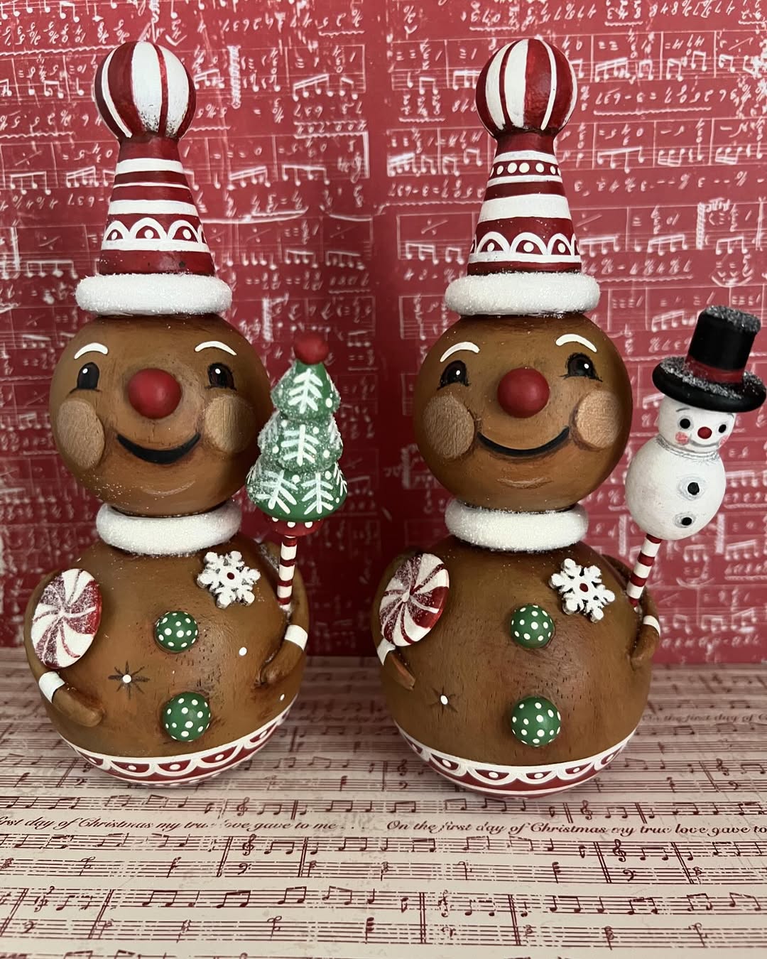 Two gingerbread figurines with red and white hats stand side by side; one holds a green Christmas tree, the other a snowman. Set against a red musical notes background, this charming Gingerbread Christmas decor adds festive cheer.