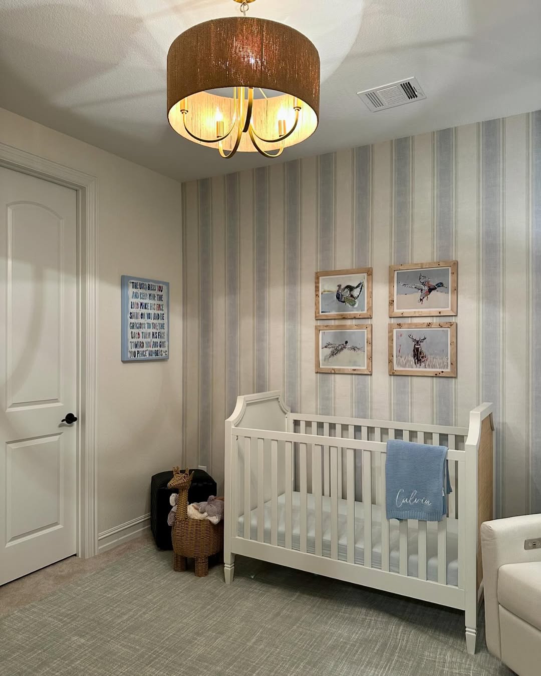 A cozy baby boy nursery featuring a white crib, wall art, blue blanket, wicker llama, and light-striped walls—perfect nursery ideas 2025 for any first-time mom.