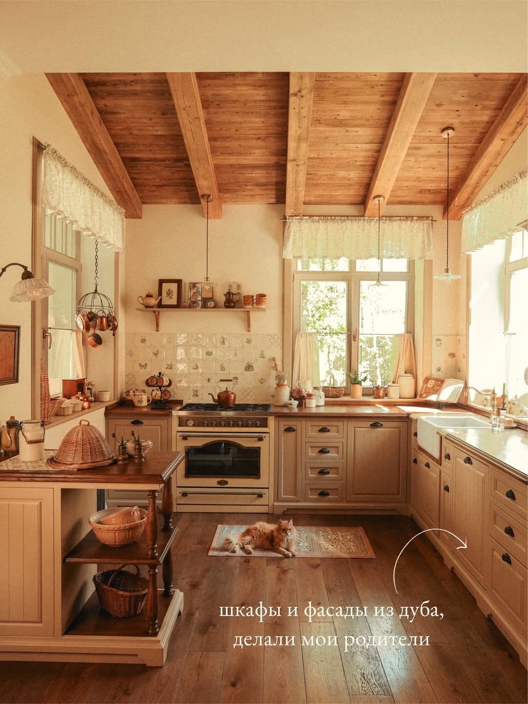 A cozy, rustic cottagecore kitchen with wooden beams, cream cabinets, large windows, and a cat lying on a rug; Russian text appears at the bottom.