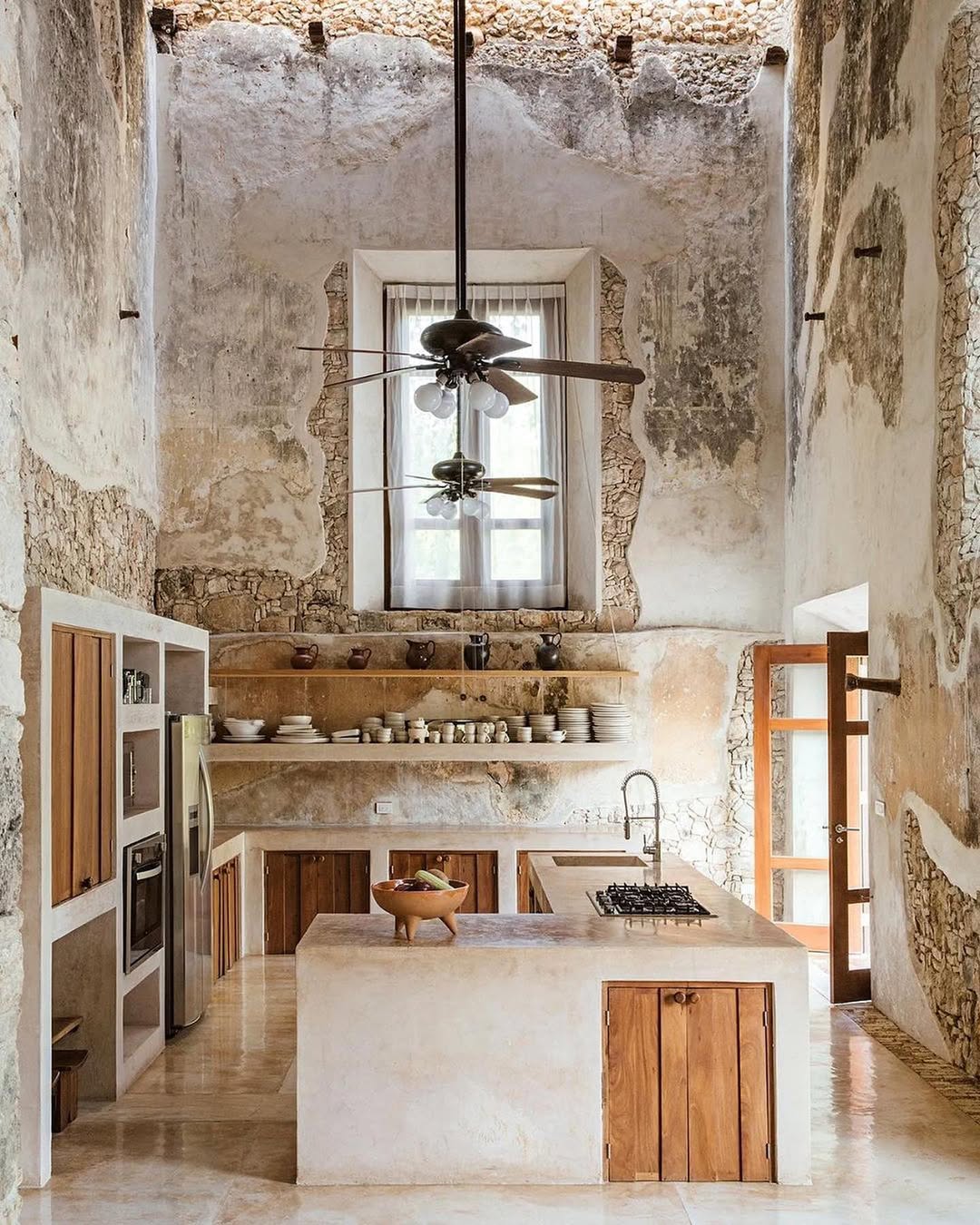 Rustic Mexican hacienda kitchen with high ceilings, exposed stone walls, open shelves displaying dishes, wood cabinets, a ceiling fan, and natural light streaming through a window and glass door.