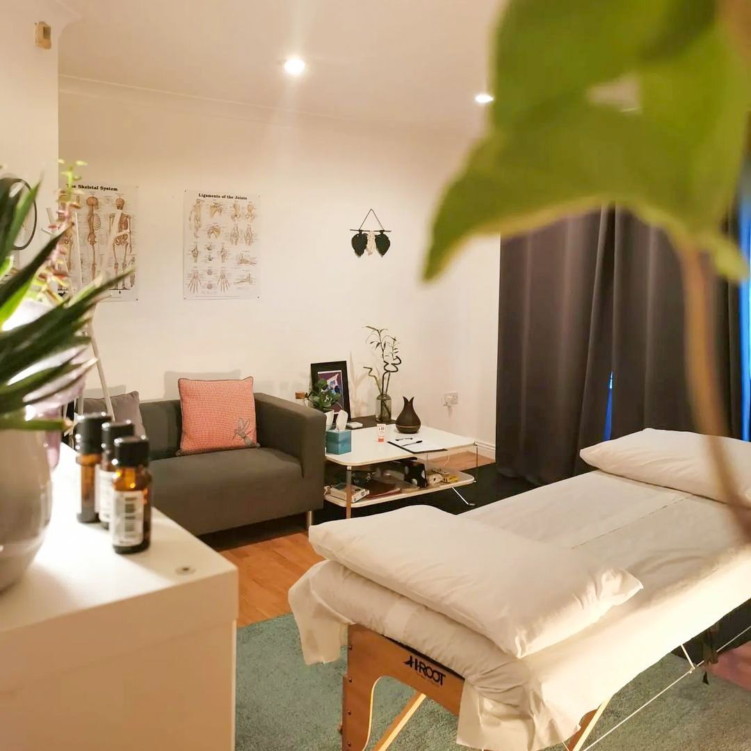 A massage therapy room featuring a treatment table, gray sofa, anatomical charts on the wall, essential oils on a counter, and indoor plants—perfect for those seeking inspiring massage room ideas.