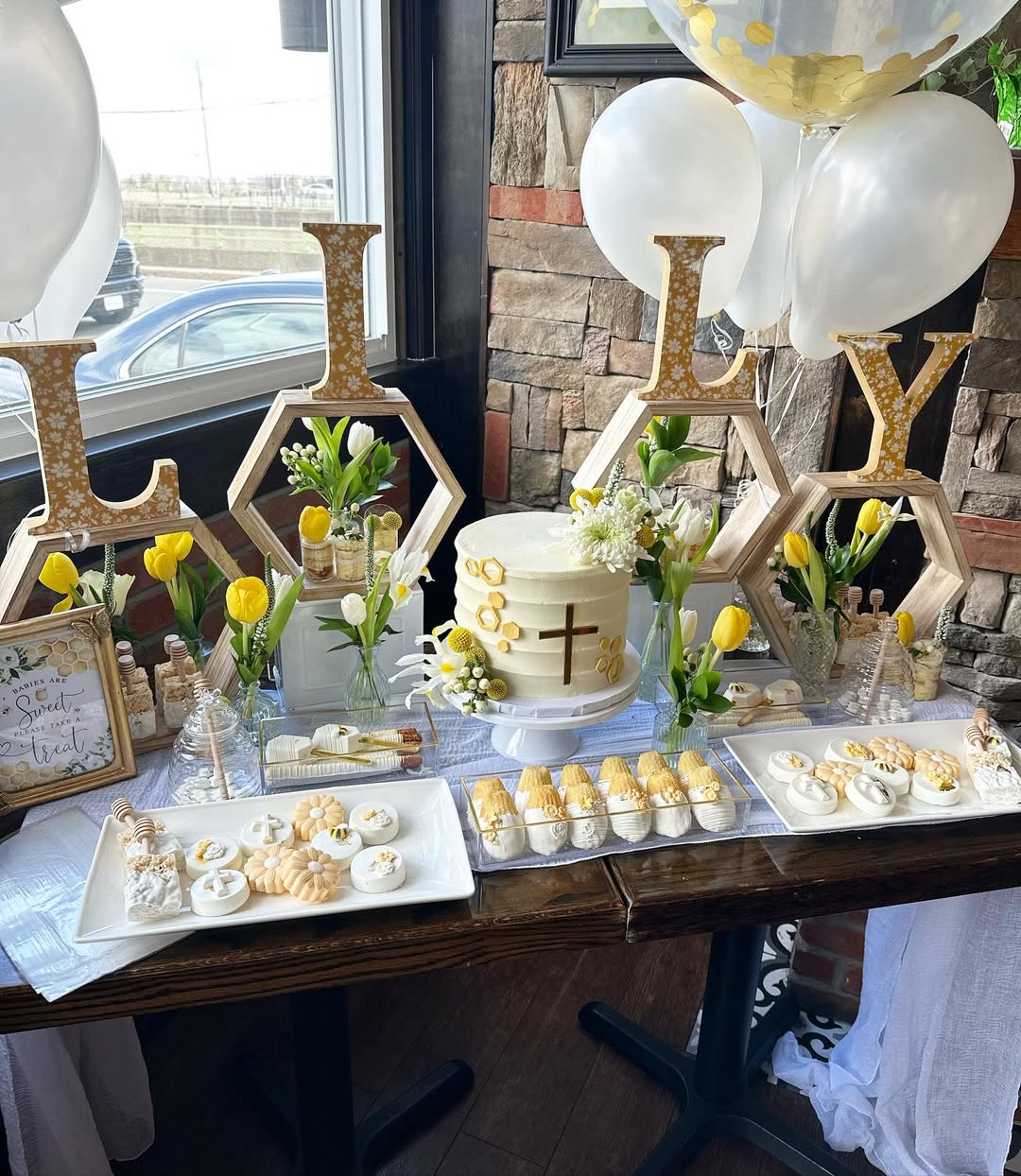 A dessert table setup with a cake, cookies, and pastries, decorated with yellow tulips, balloons, and large letters spelling