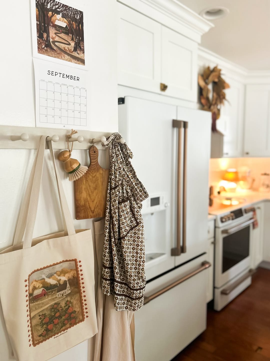 A white cottagecore kitchen with a refrigerator, stove, and September wall calendar features charming kitchen decor—a peg rack holding bags, brushes, a patterned apron, and lovely autumn decorations for cozy cottagecore kitchen ideas.