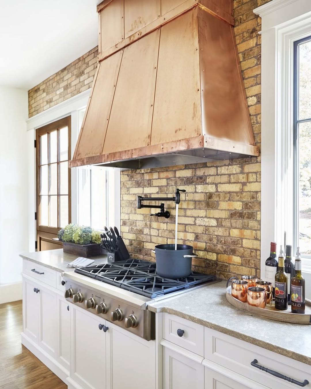 A stunning kitchen with a brick backsplash, copper range hood, gas stove, and pot filler highlights kitchen trends 2025. The countertop displays bottles, cups, and a knife block, blending timeless charm with modern style.