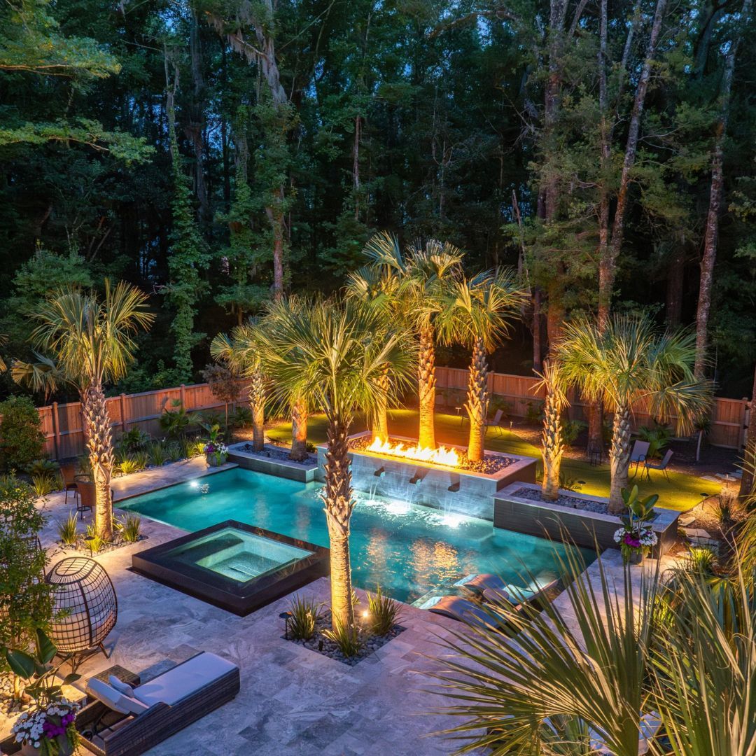 A landscaped backyard features a lit swimming pool with a built-in fire feature, surrounded by palm trees, a hot tub, lounge chairs, and seating areas at dusk—perfect inspiration for cool pool ideas.