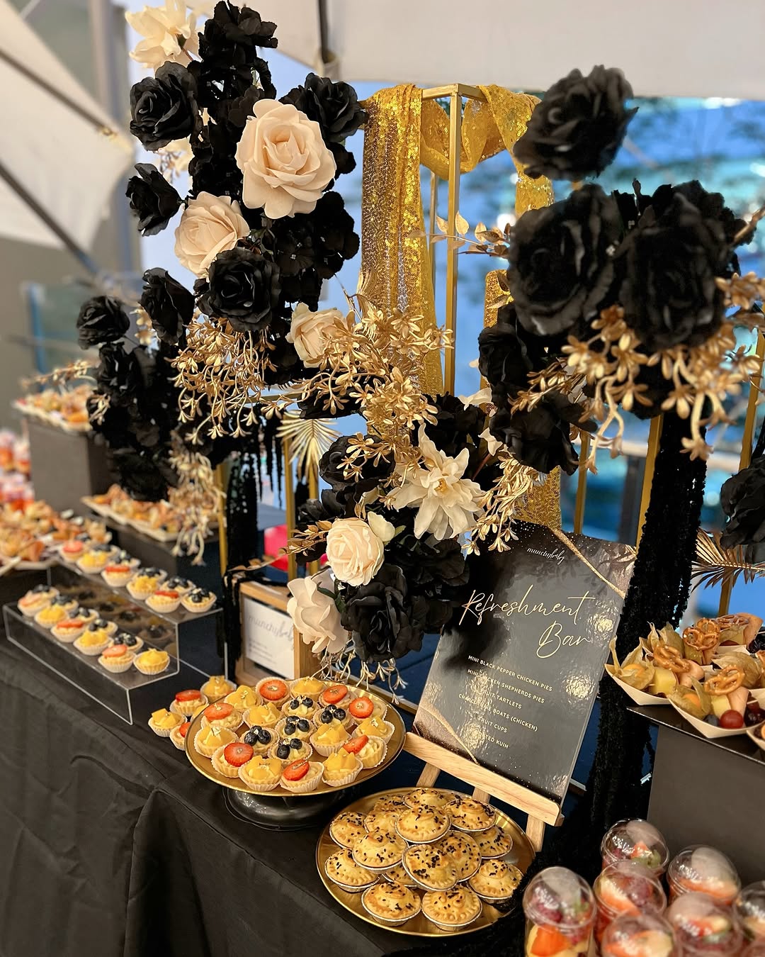 A dessert table adorned with black, white, and gold flowers showcases creative dessert table ideas, featuring assorted pastries, tarts, and a transparent sign on a sleek black tablecloth.