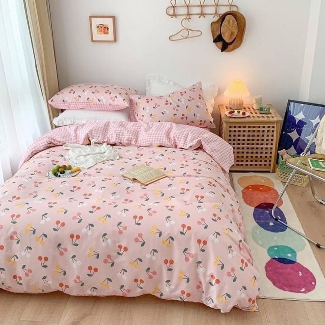 A neatly made bed with pink cherry-patterned bedding brightens this cozy room—perfect for cherry bedroom ideas. A small nightstand, a colorful rug, and hats hanging on the wall complete the cheerful look.
