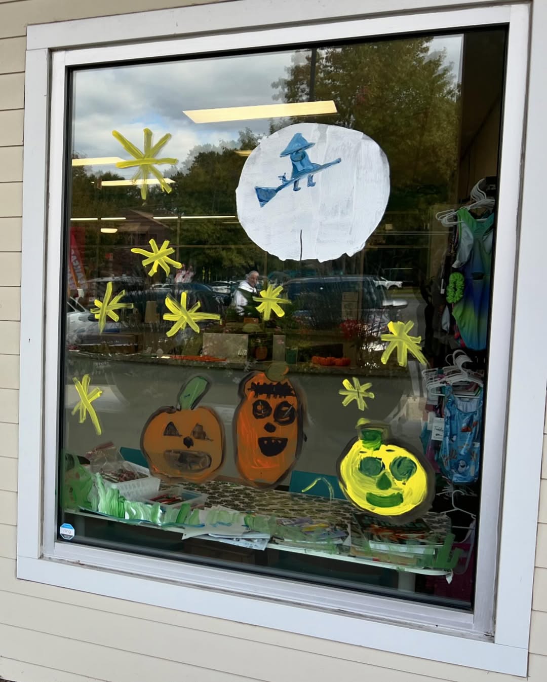A store window decorated with painted Halloween images, including pumpkins, a green face, yellow stars, and a witch flying in front of a large white moon—perfect inspiration for creative Halloween window decoration.