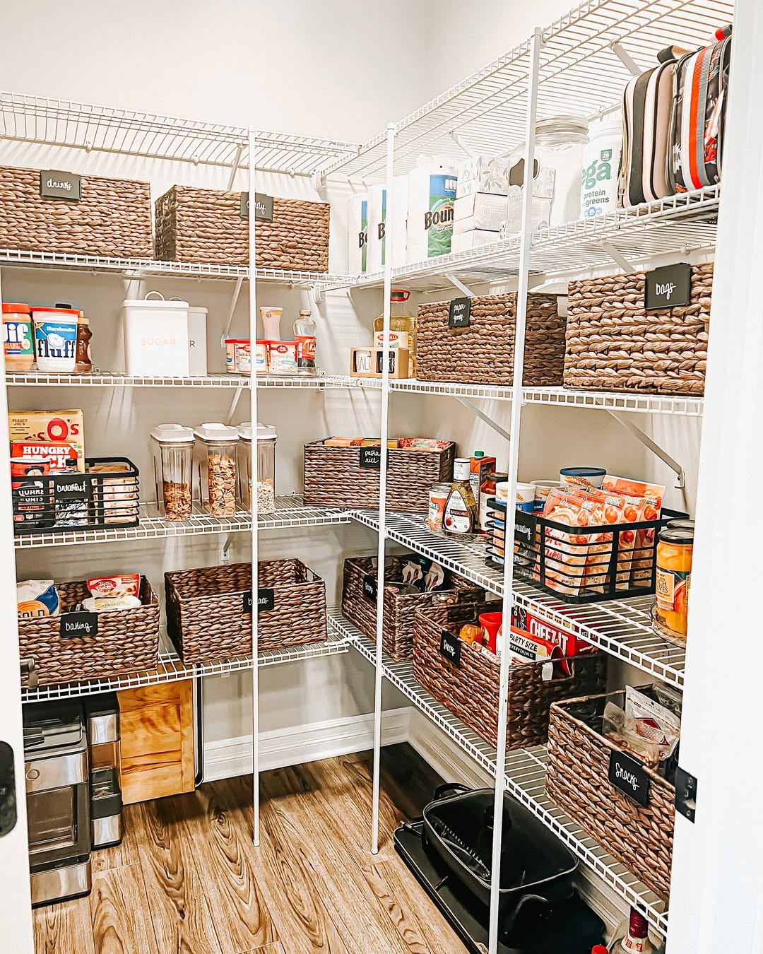 A neatly organized pantry showcases clever pantry organization with wicker baskets, labeled containers, and various packaged food items on crisp white wire shelves.