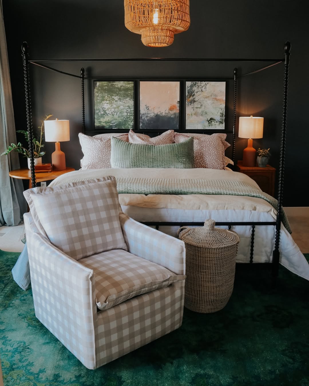 A dark feminine bedroom featuring a four-poster bed with neutral bedding, two bedside tables with lamps, a checkered armchair, a wicker basket, and a green rug on the floor.