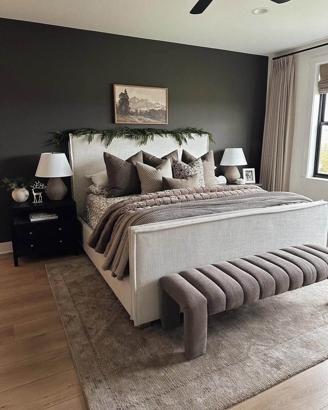 A modern gray bedroom featuring a beige upholstered bed, taupe bedding, a dark accent wall, two black nightstands with lamps, a cushioned bench, and a large window with curtains.