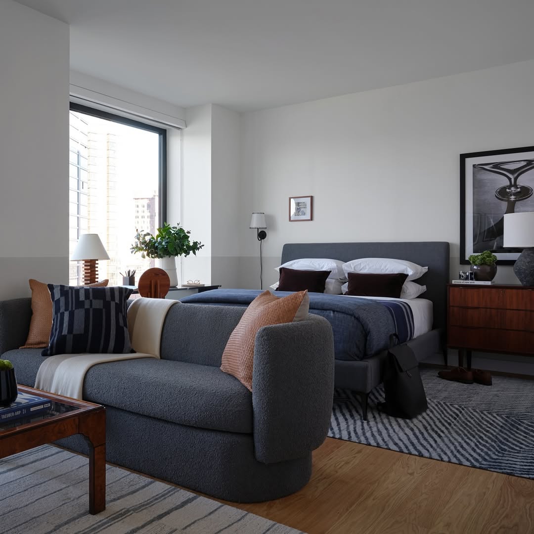 Modern bedroom in a studio apartment with a gray upholstered bed, wooden nightstand, gray sofa, coffee table, and large window for natural light—perfect for small apartment ideas. Decor features cushions, lamps, and framed art.