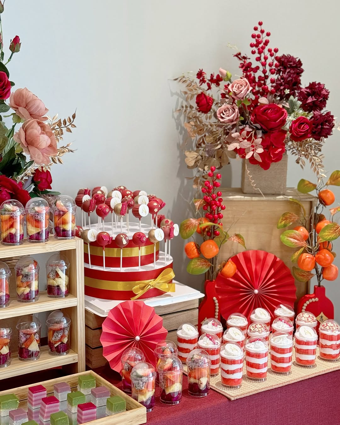A dessert table with red and white layered treats, cake pops, jelly cubes, and cupcakes—party desserts displayed among red and orange paper fans and floral arrangements in red, pink, and orange tones.