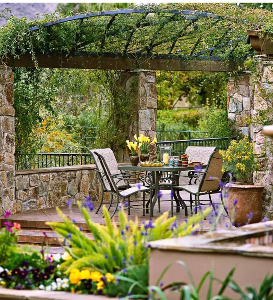 Charming small patio with a stone pergola, vines overhead, a dining table set for four, and surrounding colorful flowers and greenery.