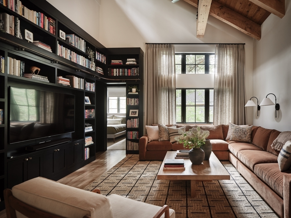 A cozy living room with a brown sectional sofa, wooden coffee table, built-in shelves, black bookshelves, large windows with curtains, and a patterned rug under a vaulted ceiling with exposed beams.
