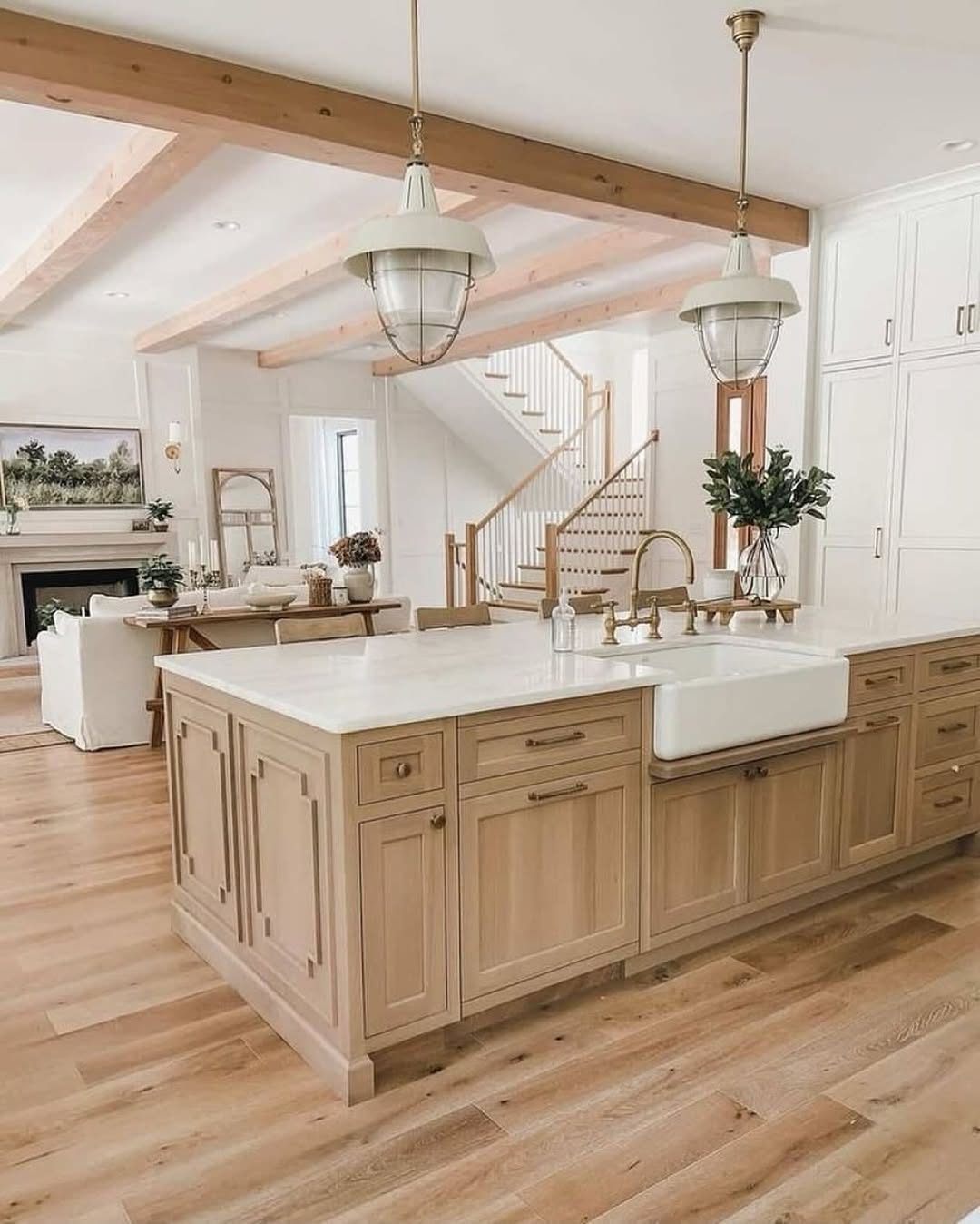 Spacious modern farmhouse kitchen with light wood cabinets, a white farmhouse sink, marble countertops, pendant lights, and open view to a living area and staircase—perfect for inspiring new kitchen ideas.