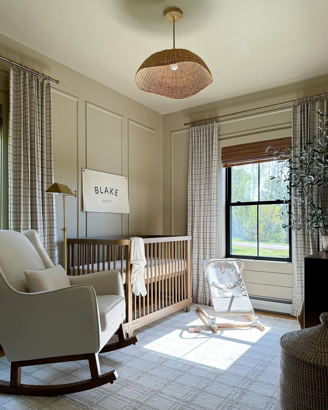 A modern baby boy nursery with a wooden crib, baby bouncer, white rocking chair, floor lamp, and large window with plaid curtains—perfect inspiration for first-time moms seeking 2025 nursery ideas. A sign above the crib reads
