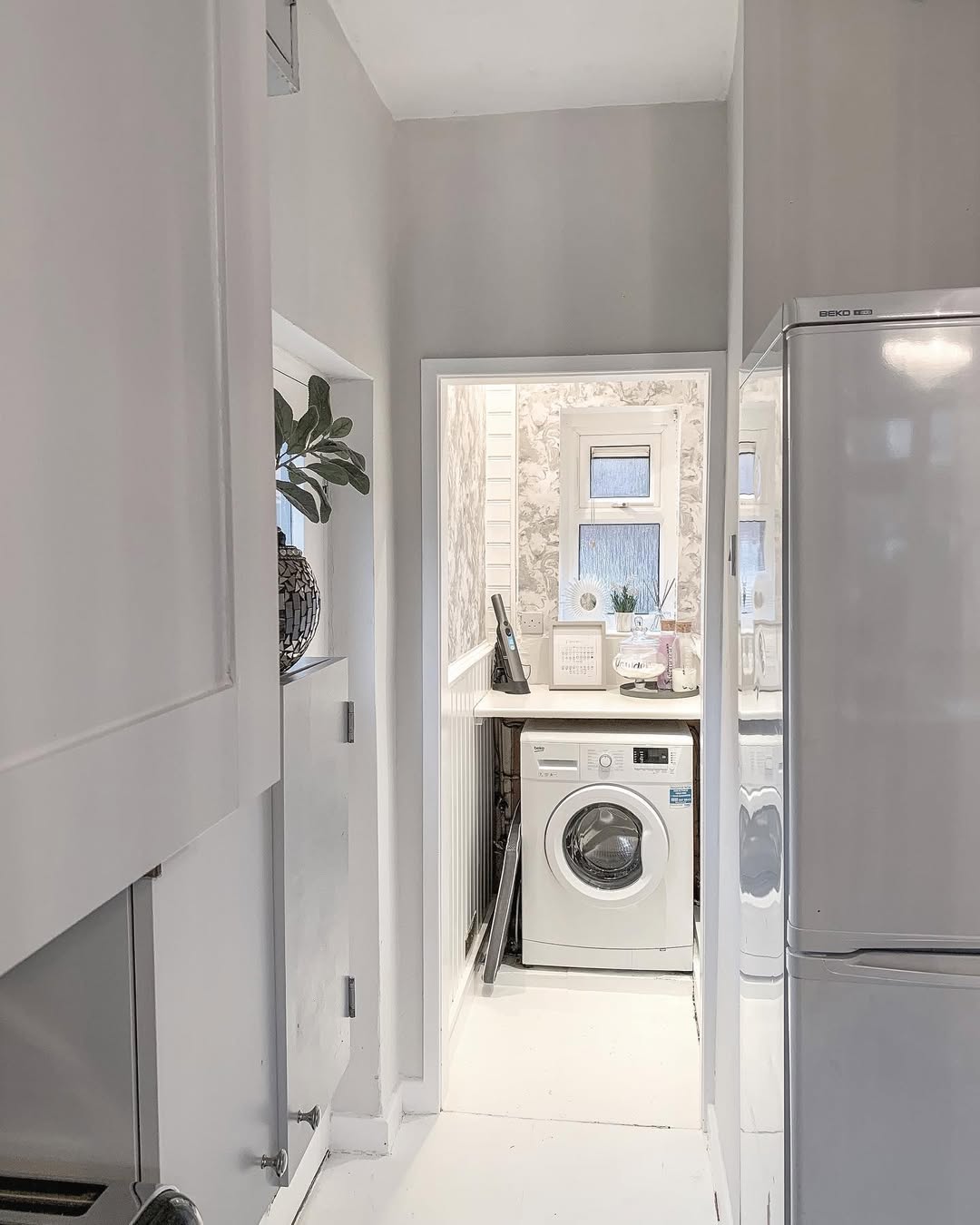 A narrow, white-walled small laundry room features a front-loading washing machine, a small window, and decorative items on the shelf above—perfect inspiration for clever laundry room design.