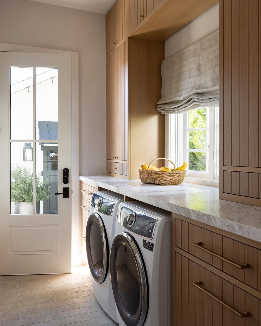 Modern laundry room with stylish laundry room cabinets, marble countertop, front-load washer and dryer, large window with Roman shade, and a basket of bananas on the counter.
