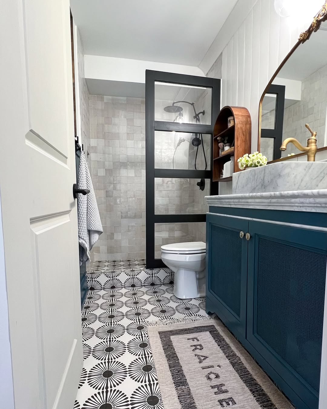 Modern bathroom design with patterned tile floor, blue vanity with gold faucet, glass shower door, towel on hook, and a shelf with decor items—perfect for inspiring new bathroom tile ideas.