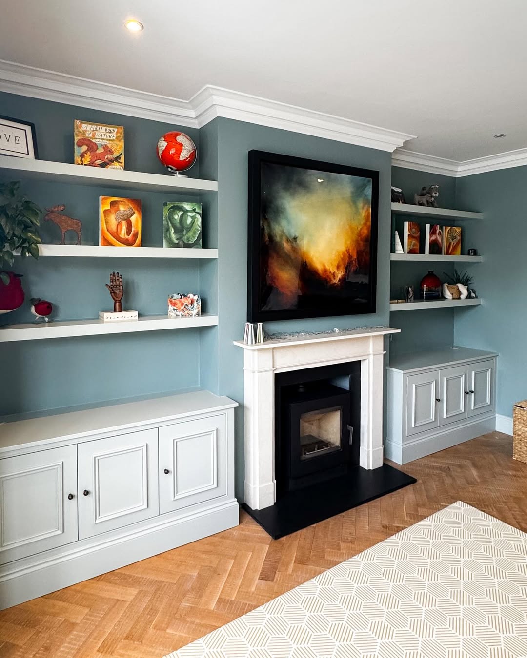 A living room with blue walls, built-in shelves for home organization holding books and decor, a white fireplace, and a large abstract painting above the mantel.