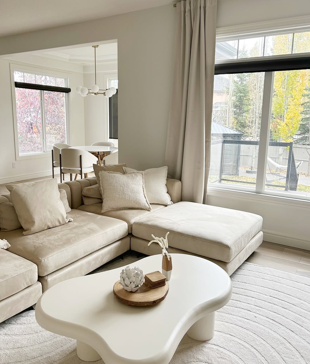 A modern, expensive looking living room with a beige sectional sofa, white coffee table, neutral decor, and large windows flooding the space with natural light—perfect for those seeking organic modern living room ideas.