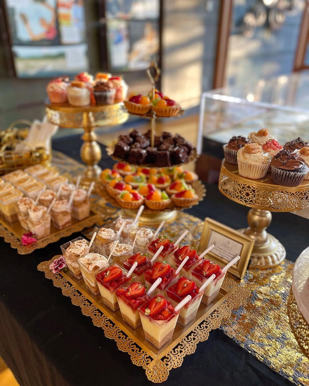 A dessert table with pretty desserts like assorted cupcakes, brownies, mini tarts, and parfait cups, all arranged on ornate gold stands and trays atop a black and gold tablecloth—perfect for inspiring your next occasion dessert table ideas.