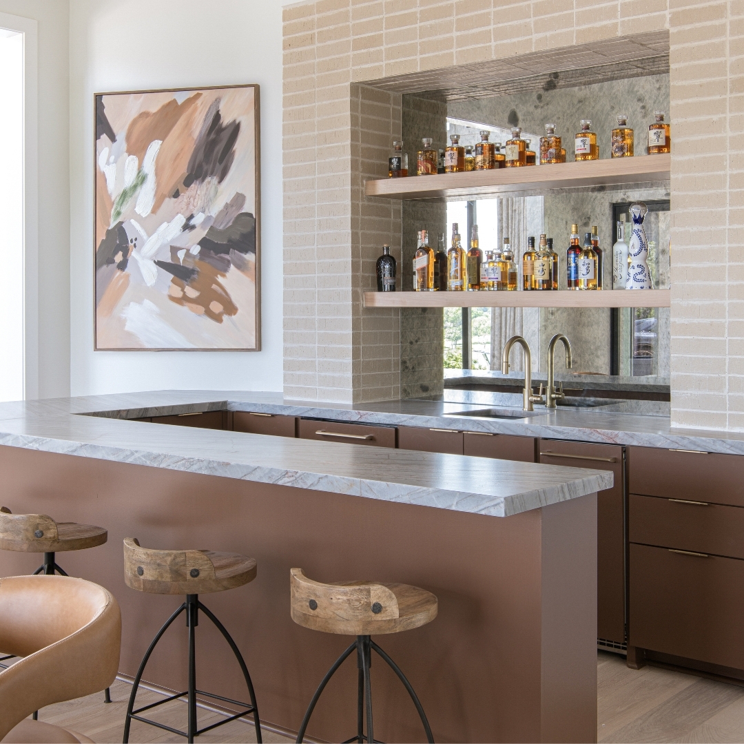 Modern home bar with a marble countertop, wooden stools, shelves displaying liquor bottles, abstract wall art, and a built-in sink—showcasing one of the latest kitchen design trends and stunning kitchen colors for a stylish space.