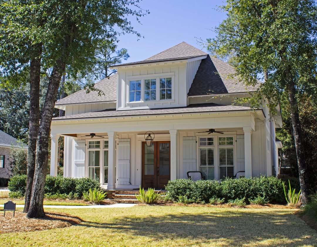 A two-story house with white siding features a coastal cottage exterior, a covered front porch, and large windows, surrounded by trees and landscaping.