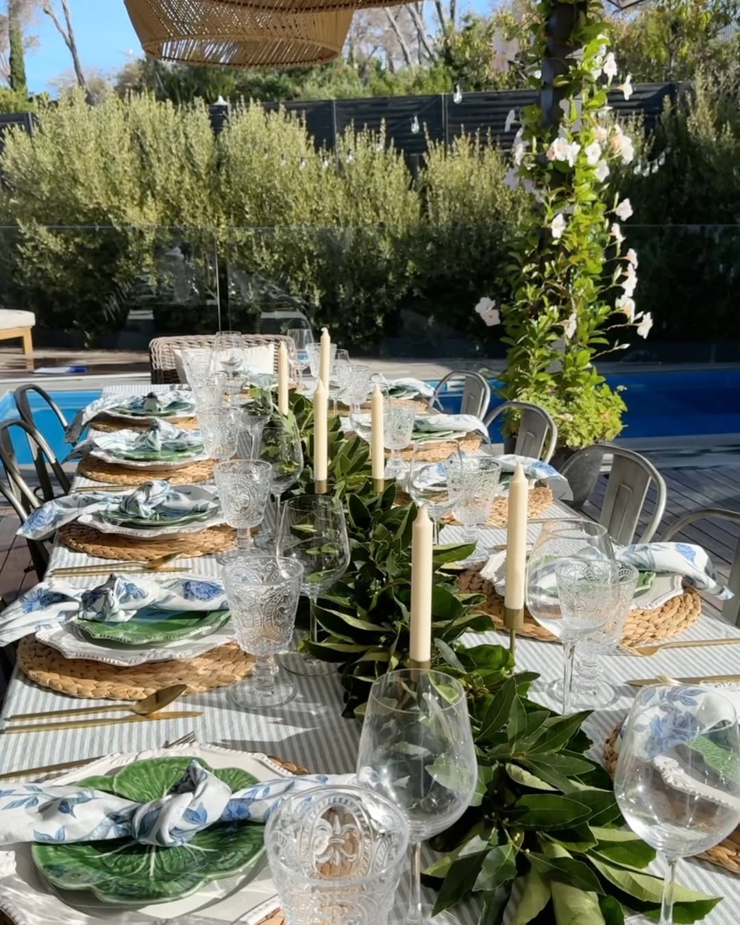 A long outdoor dining table is set with elegant table settings, including plates, napkins, glasses, candles, and a green foliage runner beside a pool and garden.