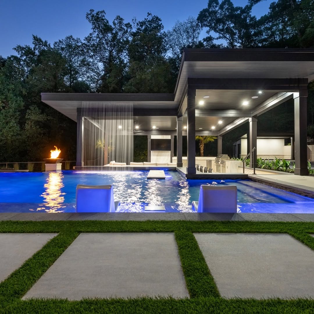 Modern outdoor pool area at dusk with glowing blue water, two submerged seats, a fire feature, overhanging roof, and surrounding trees—an inspiring example of cool pool ideas for backyard pools.