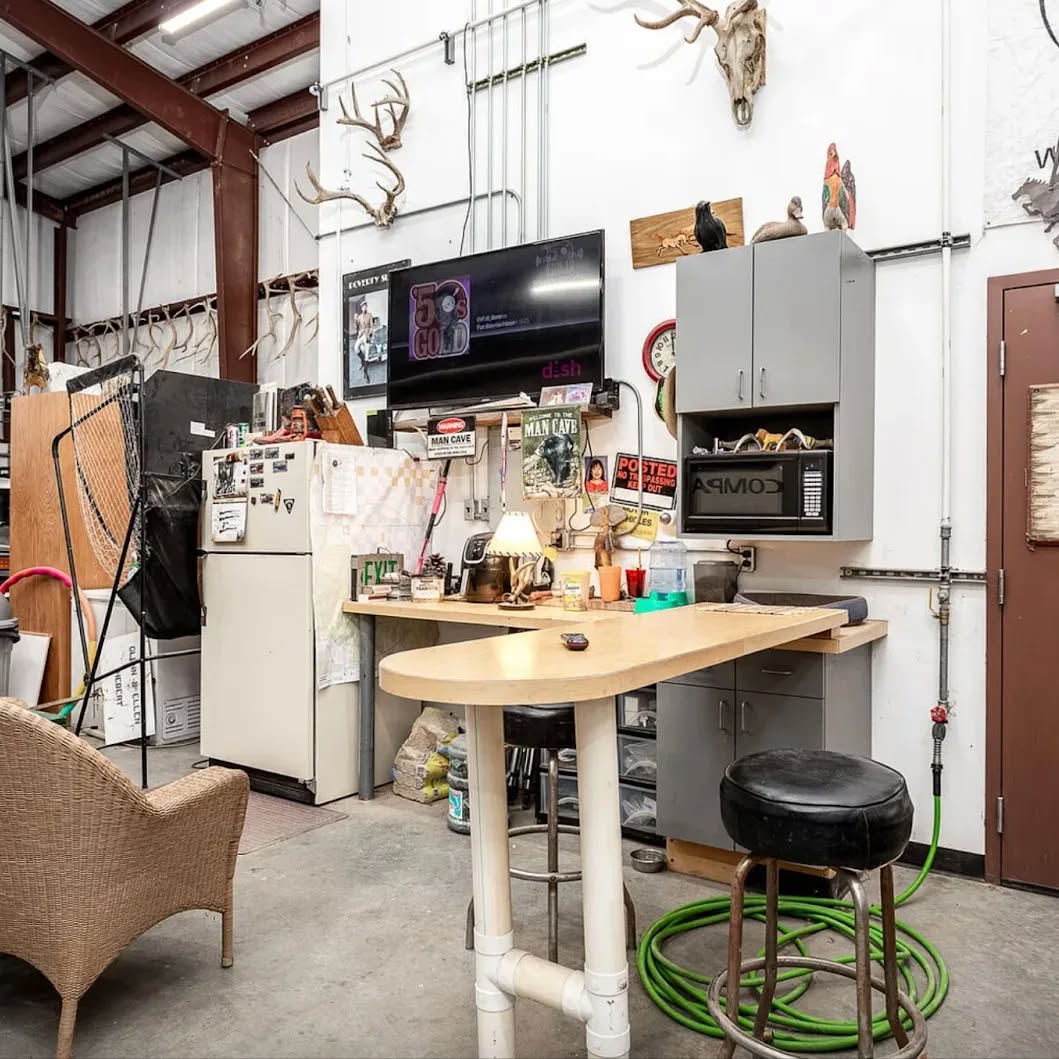 Work area in a garage or home office workshop featuring a counter, stool, fridge, mounted TV, antler decor, and various tools and supplies arranged against a white wall. Great for those seeking practical workshop ideas.